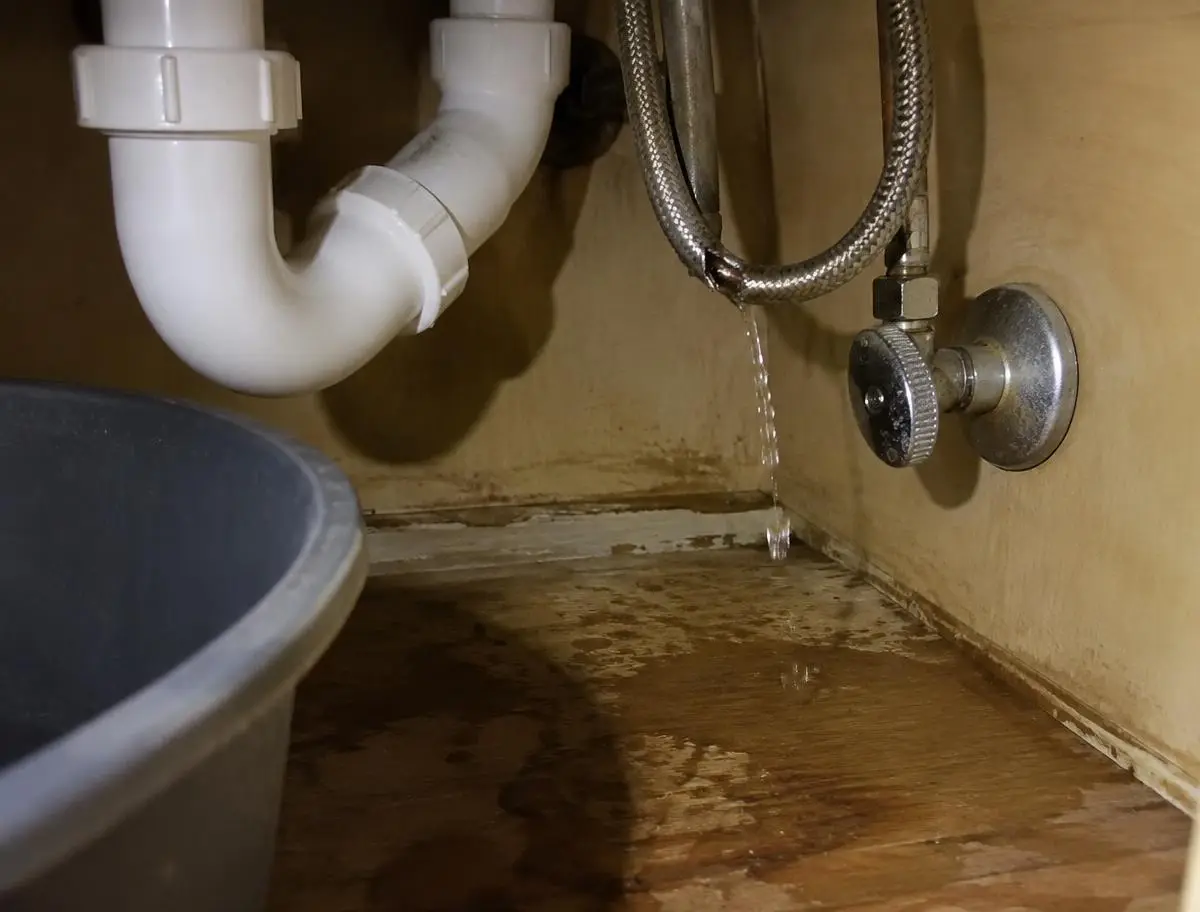 Split braided supply line dripping under a bathroom sink onto a soaked cabinet floor